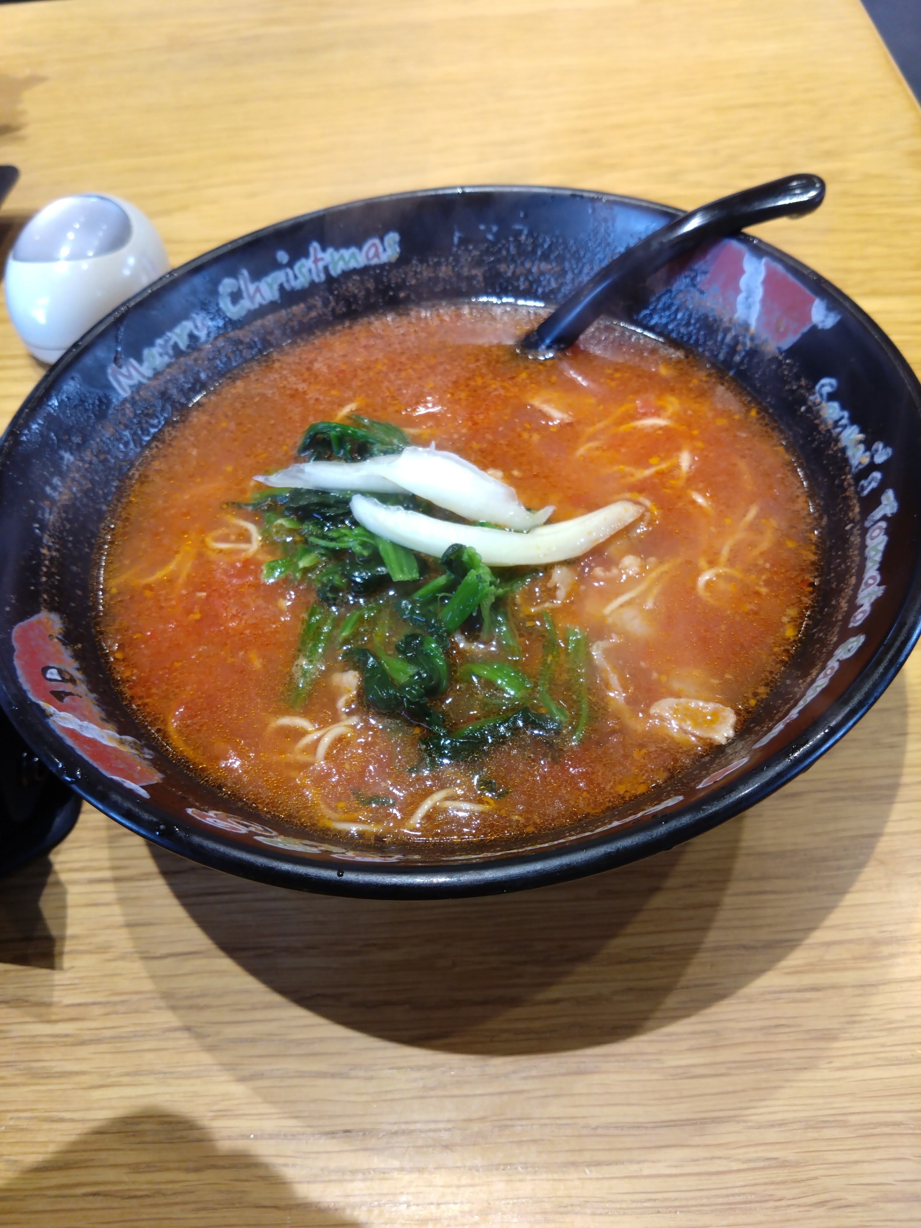 スープと麺が入った黒いラーメンの器。スープは赤く、上に青菜と白ねぎが乗っている。テーブルの上にはナプキンと小さな容器がある。